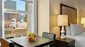 A hotel room with a bed, bedside table with a lamp, a dining table with fruit, and a view of city buildings.