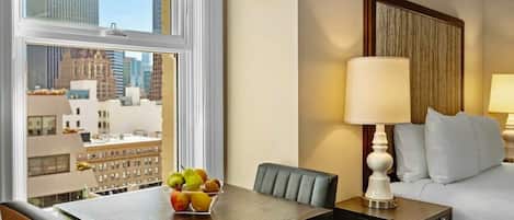 Une chambre d’hôtel avec un lit, une table de chevet avec une lampe, une table à manger avec des fruits, et une vue sur les bâtiments de la ville.