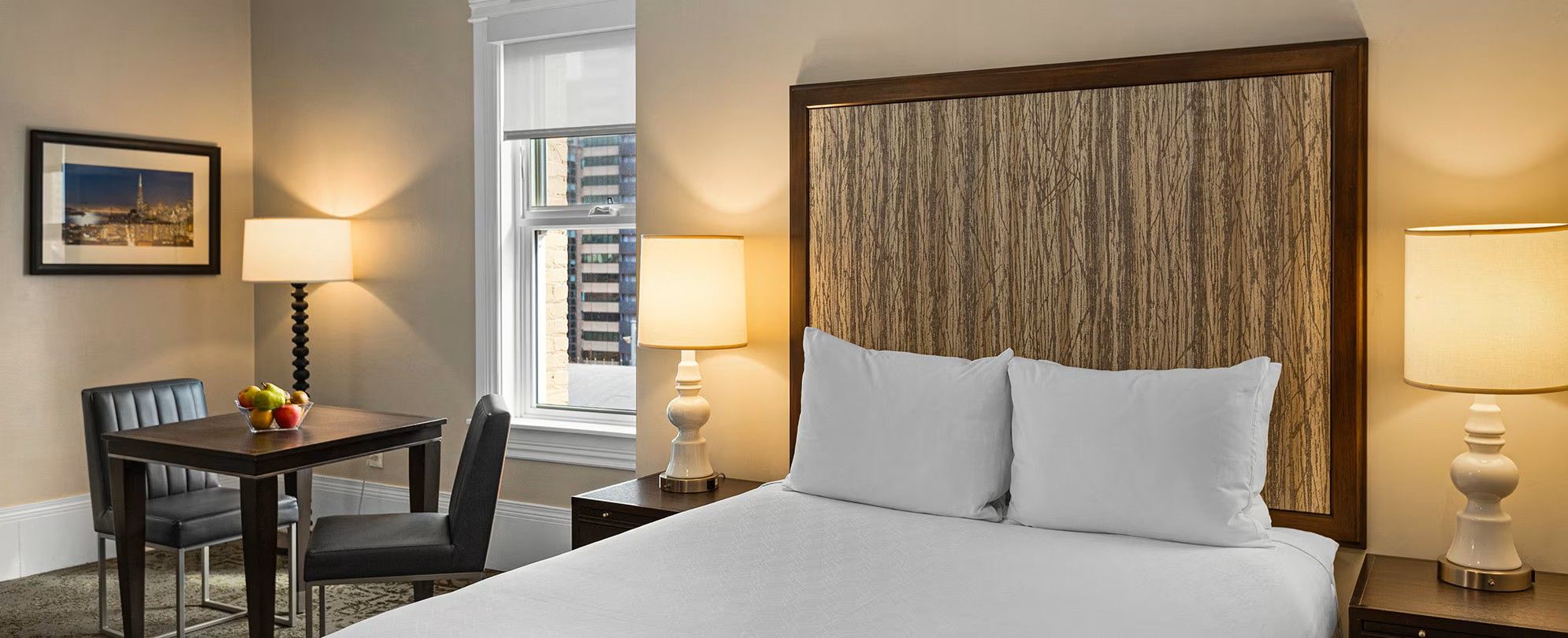 A hotel room with a bed, two bedside lamps, a desk with a chair, a fruit bowl, and a framed picture on the wall.