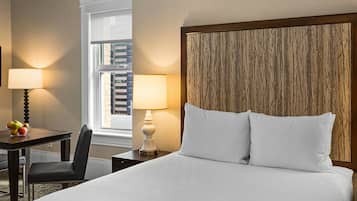 A hotel room with a bed, two bedside lamps, a desk with a chair, a fruit bowl, and a framed picture on the wall.
