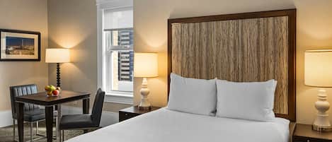 A hotel room with a bed, two bedside lamps, a desk with a chair, a fruit bowl, and a framed picture on the wall.