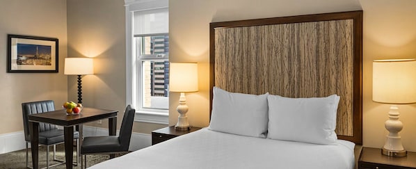 Habitación de hotel con una cama, dos lámparas de noche, un escritorio con silla, un frutero y un cuadro en la pared.