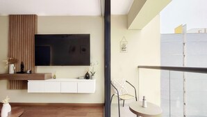 Uma sala de estar moderna com televisão montada, um armário branco pequeno, uma poltrona de vime e uma varanda com guarda-corpo de vidro.