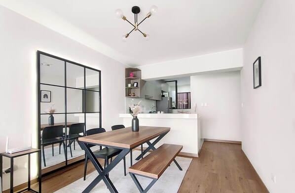 Un comedor moderno con una mesa de madera, sillas negras y un banco. Se ve una cocina al fondo, con una encimera blanca y diversos objetos.