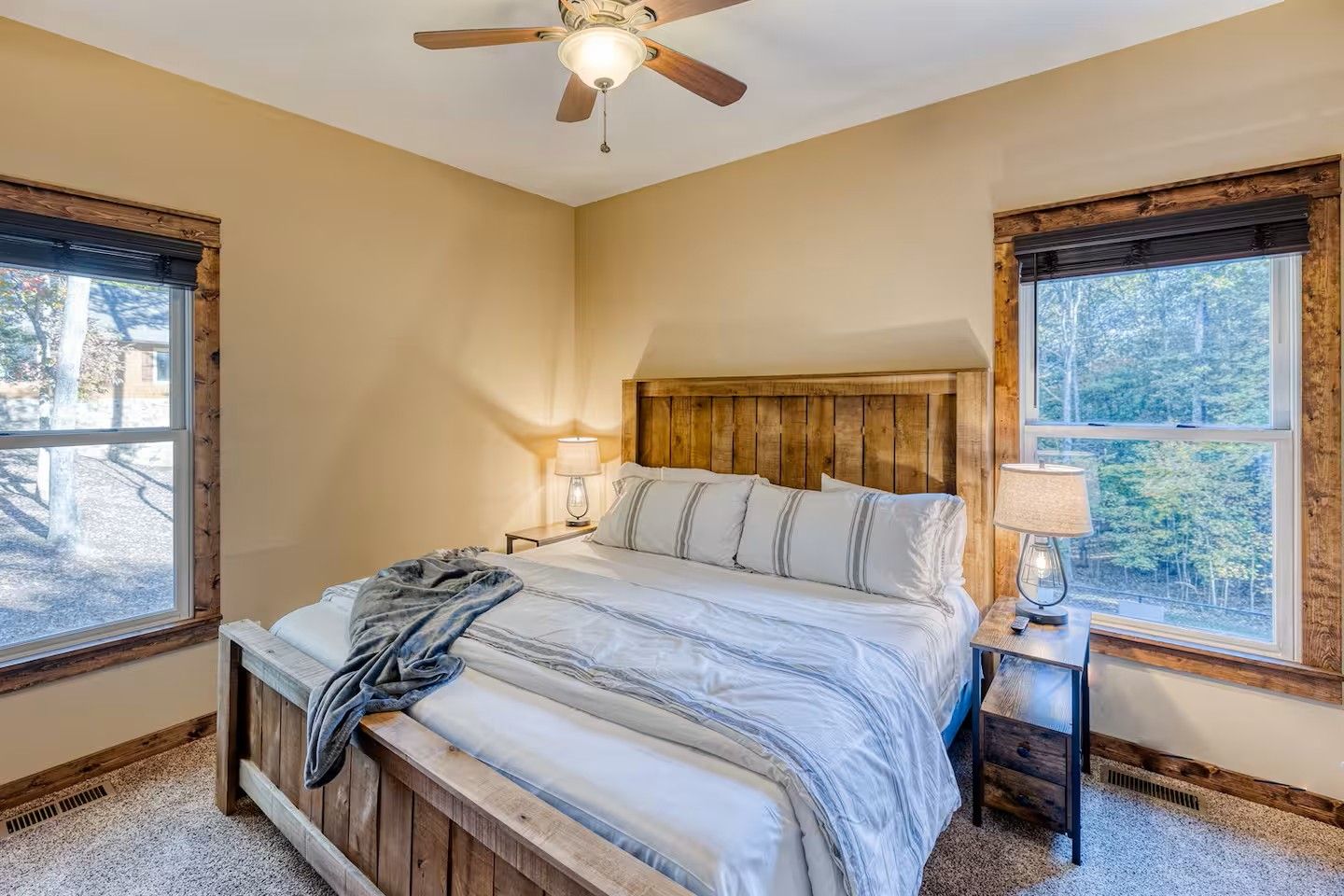 A bedroom with a bed, two bedside lamps, a ceiling fan, and windows with views of trees.