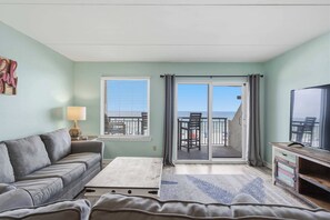 A room with a grey sofa, a small table, a TV on a wooden stand, a window with blinds, and a view of the beach through a sliding door. - Beachfront Paradise with Stunning Gulf Views, Island Echo Condo (Fort Walton Beach)