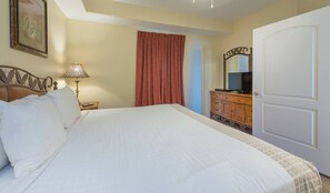 A neatly made bed with white linens, a wooden nightstand with a lamp, a television on a wooden dresser, and a large white door. - Disney Dreams Apartment Amazing Lake Views! (Orlando)