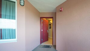Um corredor com uma porta vermelha, uma janela com cortinas, uma luminária na parede e um tapete de entrada.