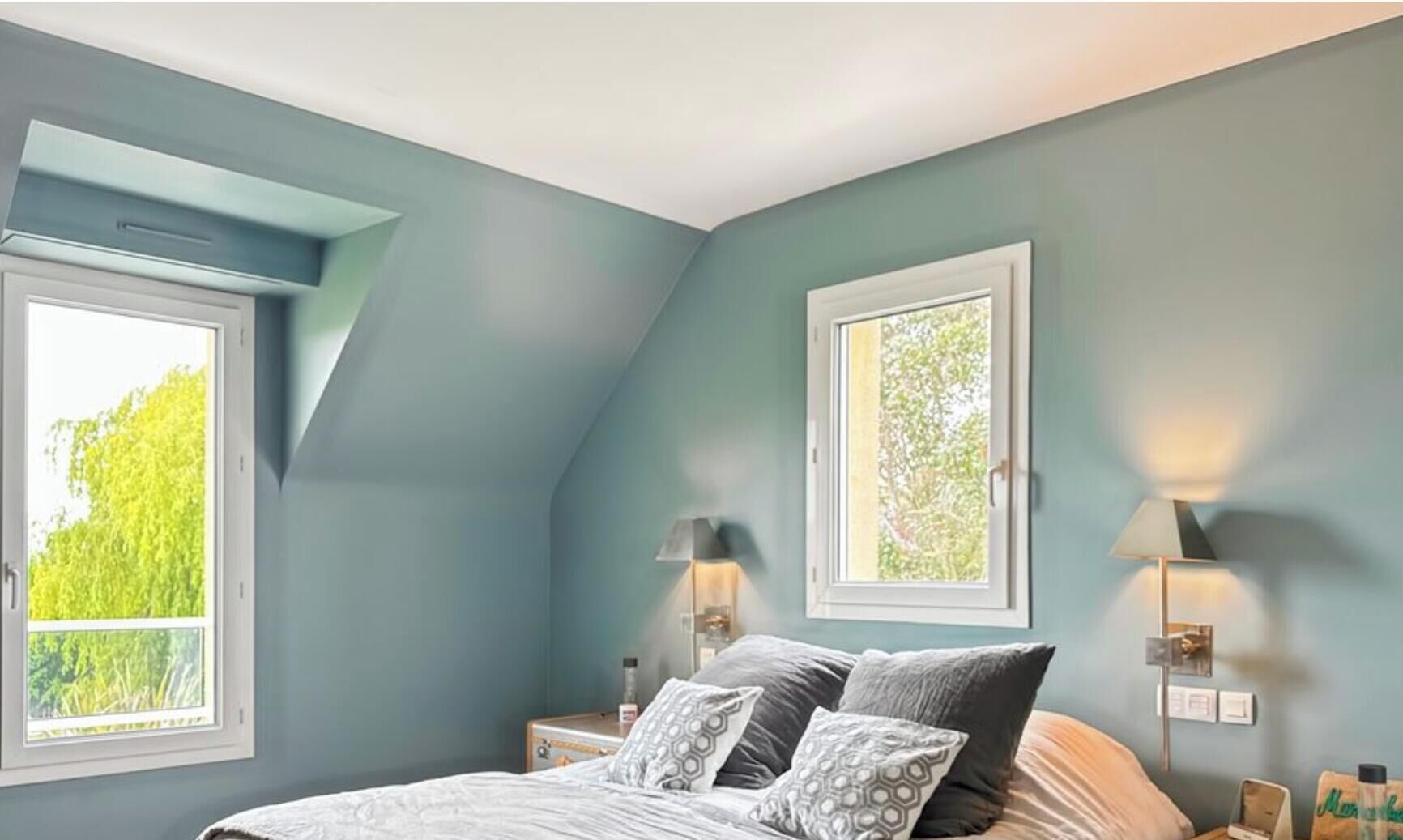 A bedroom with a sloped ceiling, two bedside lamps, a bed with patterned bedding, and two windows with white frames.