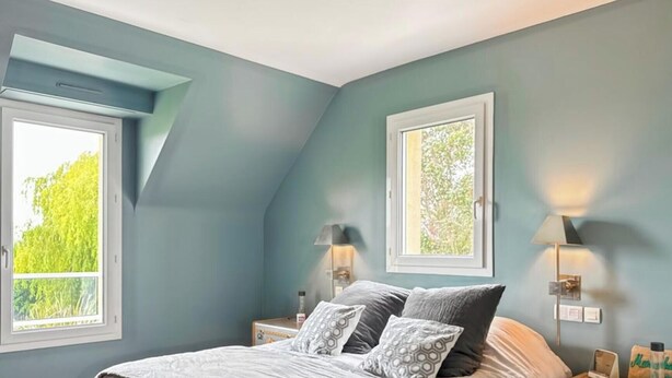 Un dormitorio con techo inclinado, dos lámparas de noche, una cama con ropa de cama estampada y dos ventanas con marcos blancos.