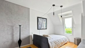 Un dormitorio moderno con cama, un cuadro, una lámpara colgante y vista a zonas verdes por una ventana.