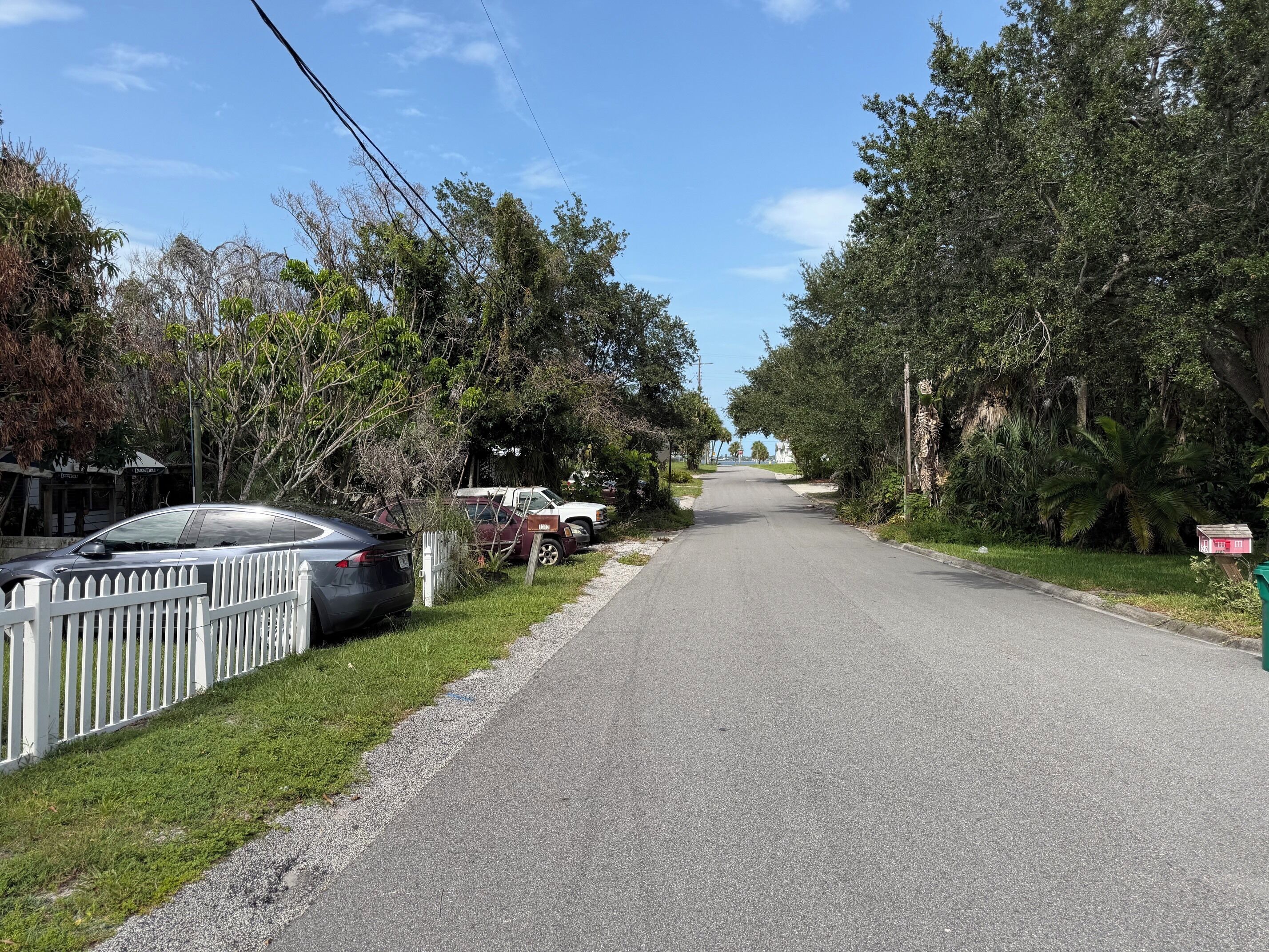 A quiet street lined with parked cars and trees, leading to a building with a sign that reads 'Hotel'.