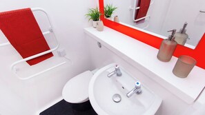 Ein Badezimmer mit einem weißen Waschbecken, zwei Wasserhähnen, einem roten Handtuch, einer kleinen Pflanze und einem Spiegel mit rotem Rahmen.