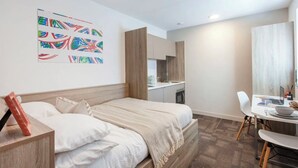 A modern hotel room with a bed, a desk, a chair, a kitchen area, and a colorful abstract painting on the wall. - Student Only Berkshire House (Reading)