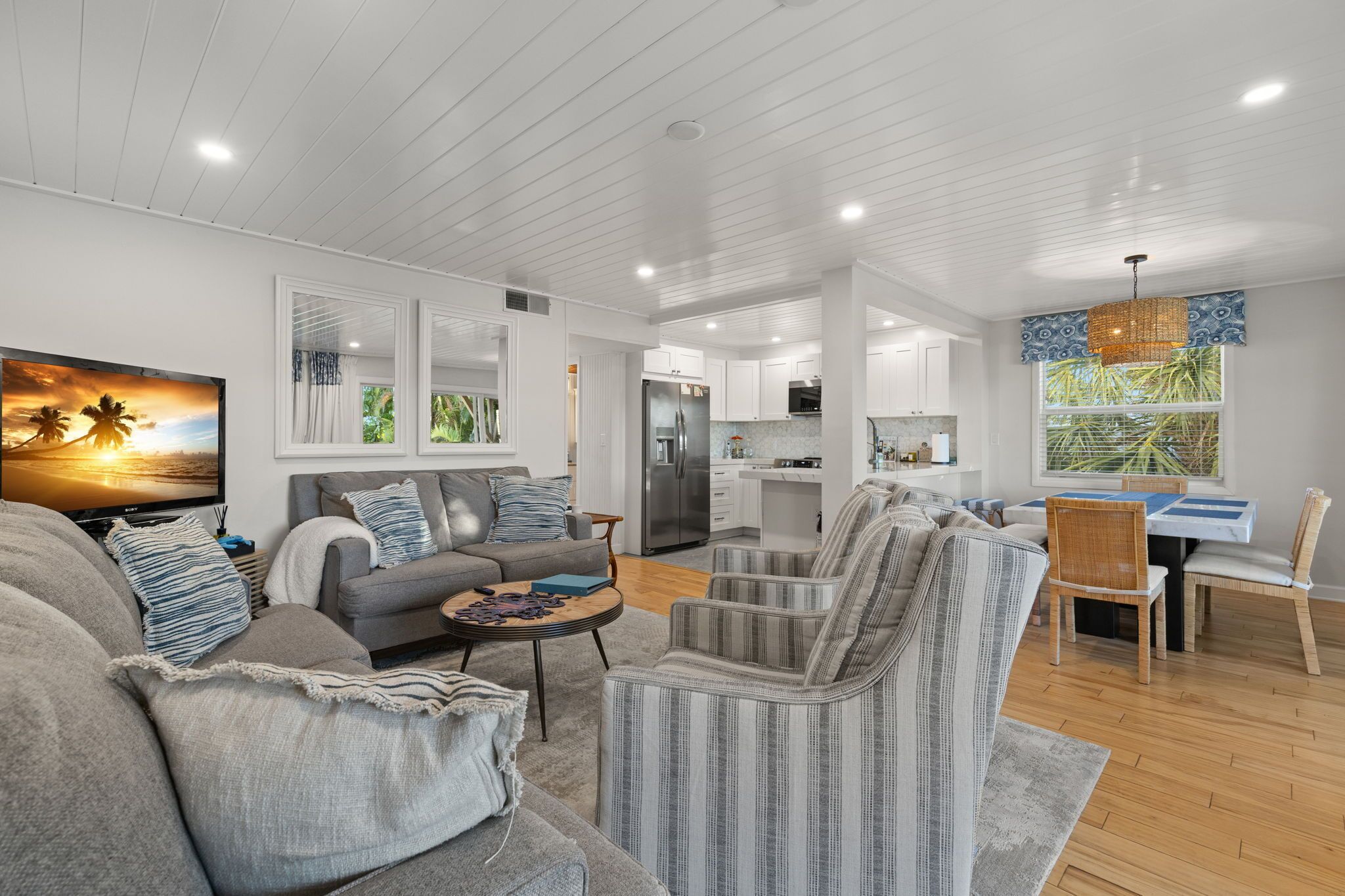 A modern living room with a sofa, armchair, and coffee table, a dining area with a table and chairs, and a kitchen with a refrigerator and stove.