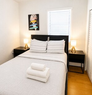 A neatly made bed with white bedding and pillows, two bedside tables with lamps, a window with blinds, and a wall-mounted picture. - Spacious Miami Beach 1BR • Beach Access • Sleeps 3 (Miami Beach)