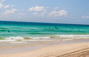 Ein Strand mit klarem türkisblauem Wasser, einem Sandufer und einigen badenden Personen.
