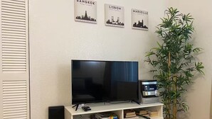 Ein Wohnzimmer mit Fernseher auf einem weißen Schrank, einem Lautsprecher, einer Pflanze und Wandbildern von Stadtansichten.