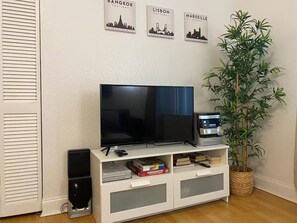 Ein Wohnzimmer mit Fernseher auf einem weißen Schrank, einem Lautsprecher, einer Pflanze und Wandbildern von Stadtansichten.
