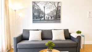 Ein modernes Wohnzimmer mit grauem Sofa, zwei weißen Kissen, einem weißen Couchtisch mit Pflanze und Dekoration, sowie einem schwarz-weißen Stadtbild an der Wand.