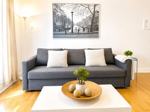 Ein modernes Wohnzimmer mit grauem Sofa, zwei weißen Kissen, einem weißen Couchtisch mit Pflanze und Dekoration, sowie einem schwarz-weißen Stadtbild an der Wand.