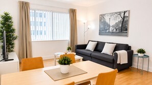 Ein modernes Wohnzimmer mit einer Couch, einem Esstisch, Stühlen, einer Topfpflanze, einer Lampe und einem gerahmten Bild an der Wand.