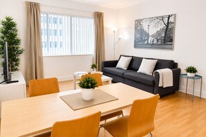Ein modernes Wohnzimmer mit einer Couch, einem Esstisch, Stühlen, einer Topfpflanze, einer Lampe und einem gerahmten Bild an der Wand.