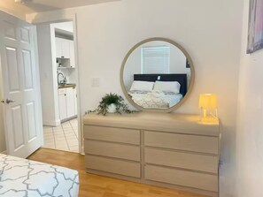 Ein Schlafzimmer mit einem Bett, einem Schminktisch, einem Spiegel, einer Nachttischlampe und einer Zimmerpflanze.