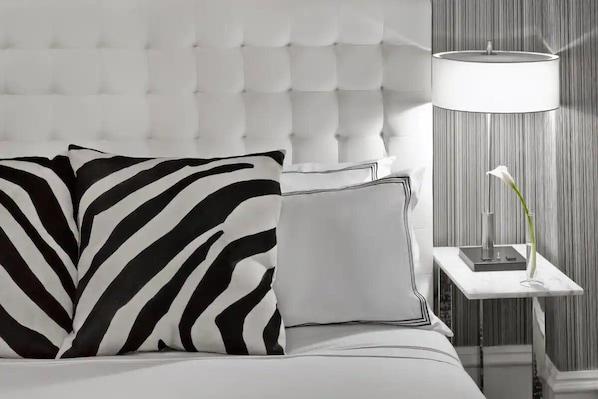 Ein Bett mit einer karopragigen Kopfteile, ein Zebra-Kissen, ein schlichtes Kissen, ein Nachttisch mit Lampe und eine Vase mit einer einzelnen Blume.