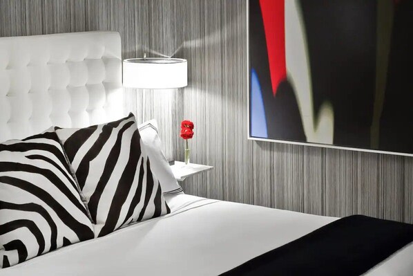 Ein Hotelzimmer mit einer gesteppten Kopfteil, einem weißen Bett mit schwarz-weiß gestreiften Kissen, einem Nachttisch mit einer Lampe und einer roten Blumenvase sowie einem großen abstrakten Gemälde an der Wand.