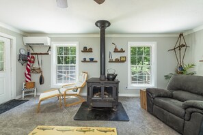 A cozy living room with a wood-burning stove, two windows, a chair, a sofa, and various decorative items. - Little Rock Creek Cottage on Trout Stream (Bakersville)