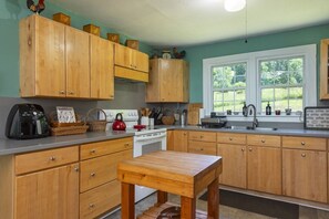 A kitchen with wooden cabinets, a stove, a sink, and a window with a view of greenery. - Little Rock Creek Cottage on Trout Stream (Bakersville)