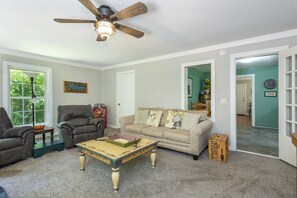 A living room with a ceiling fan, a beige sofa, two armchairs, a coffee table, and a window with a view of trees. - Little Rock Creek Cottage on Trout Stream (Bakersville)