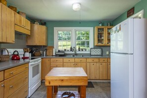 A kitchen with wooden cabinets, a white refrigerator, and a window with a view of greenery. - Little Rock Creek Cottage on Trout Stream (Bakersville)