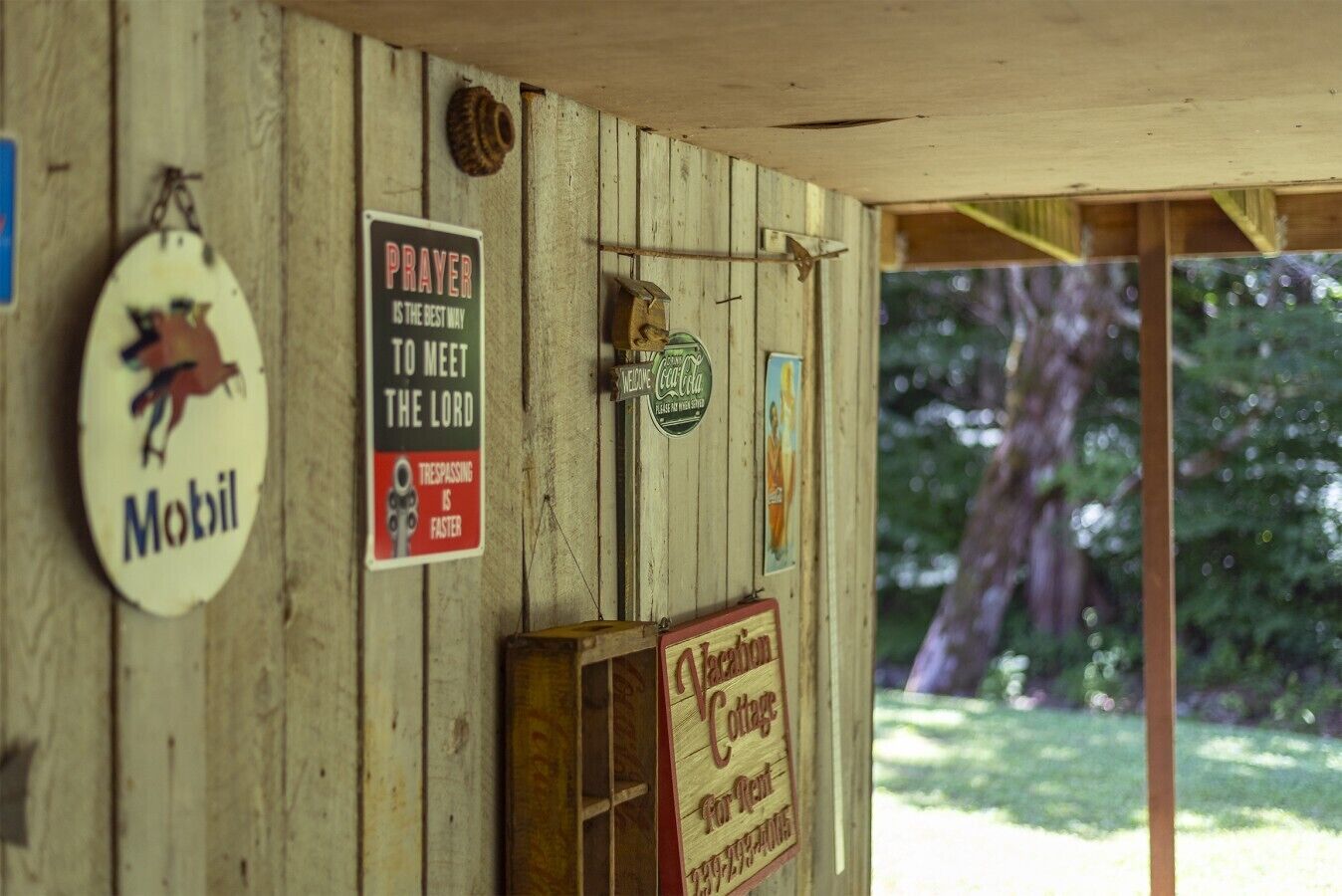 A wooden cabin with a sign for Mobil, a sign stating 'Prayer is the best way to meet the Lord,' a Coca-Cola sign, and a 'Welcome to Cedar Cottage For Rent' sign.