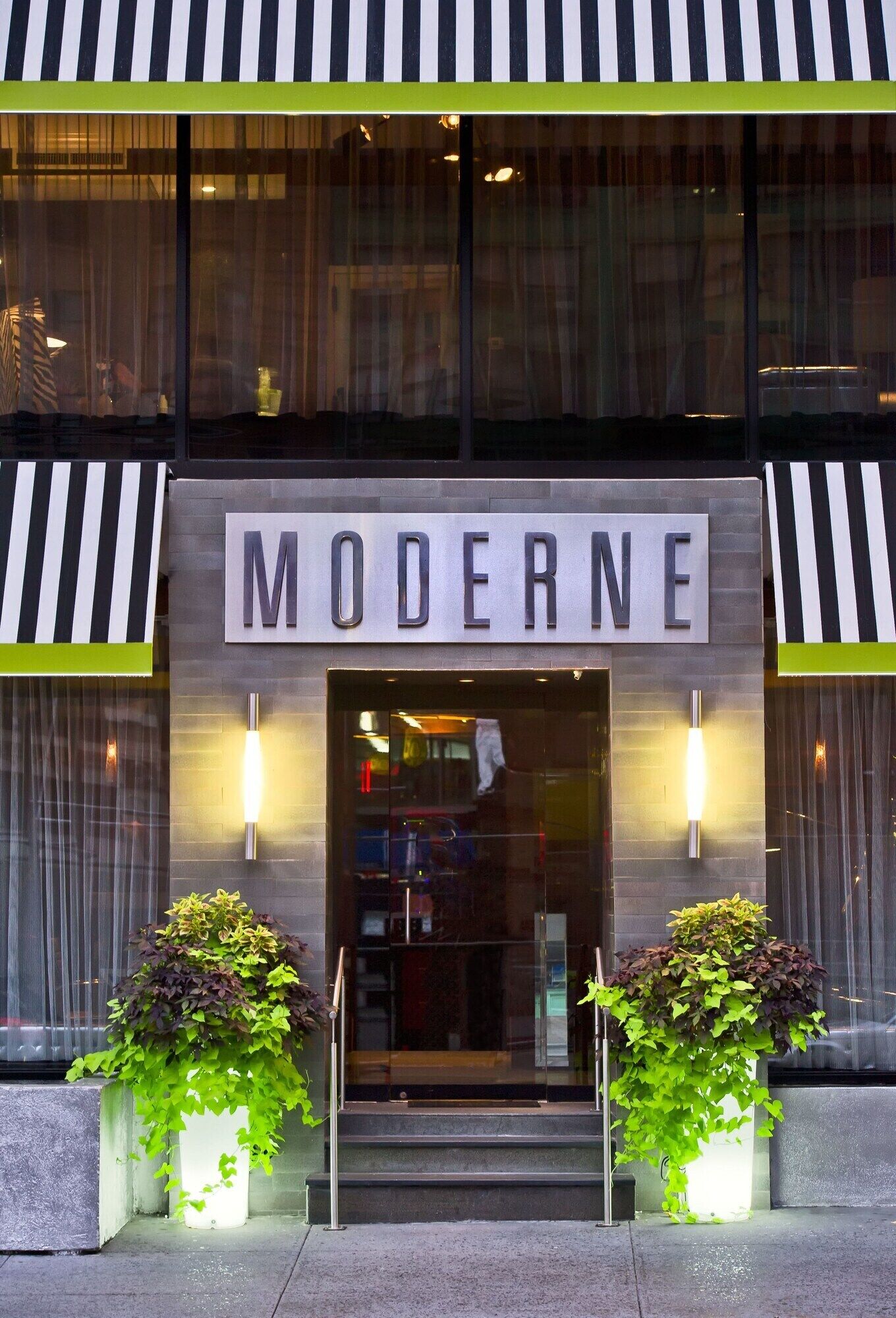 Um edifício com uma placa indicando 'MODERNE' acima da entrada, duas plantas em vasos de cada lado e estilo arquitetônico moderno.
