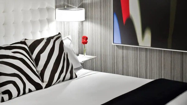 Ein Hotelzimmer mit einer gesteppten Kopfteil, einem weißen Bett mit schwarz-weiß gestreiften Kissen, einem Nachttisch mit einer Lampe und einer roten Blumenvase sowie einem großen abstrakten Gemälde an der Wand.