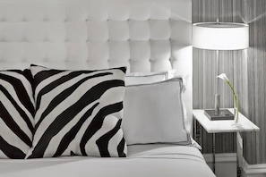 Ein Bett mit einer karopragigen Kopfteile, ein Zebra-Kissen, ein schlichtes Kissen, ein Nachttisch mit Lampe und eine Vase mit einer einzelnen Blume.