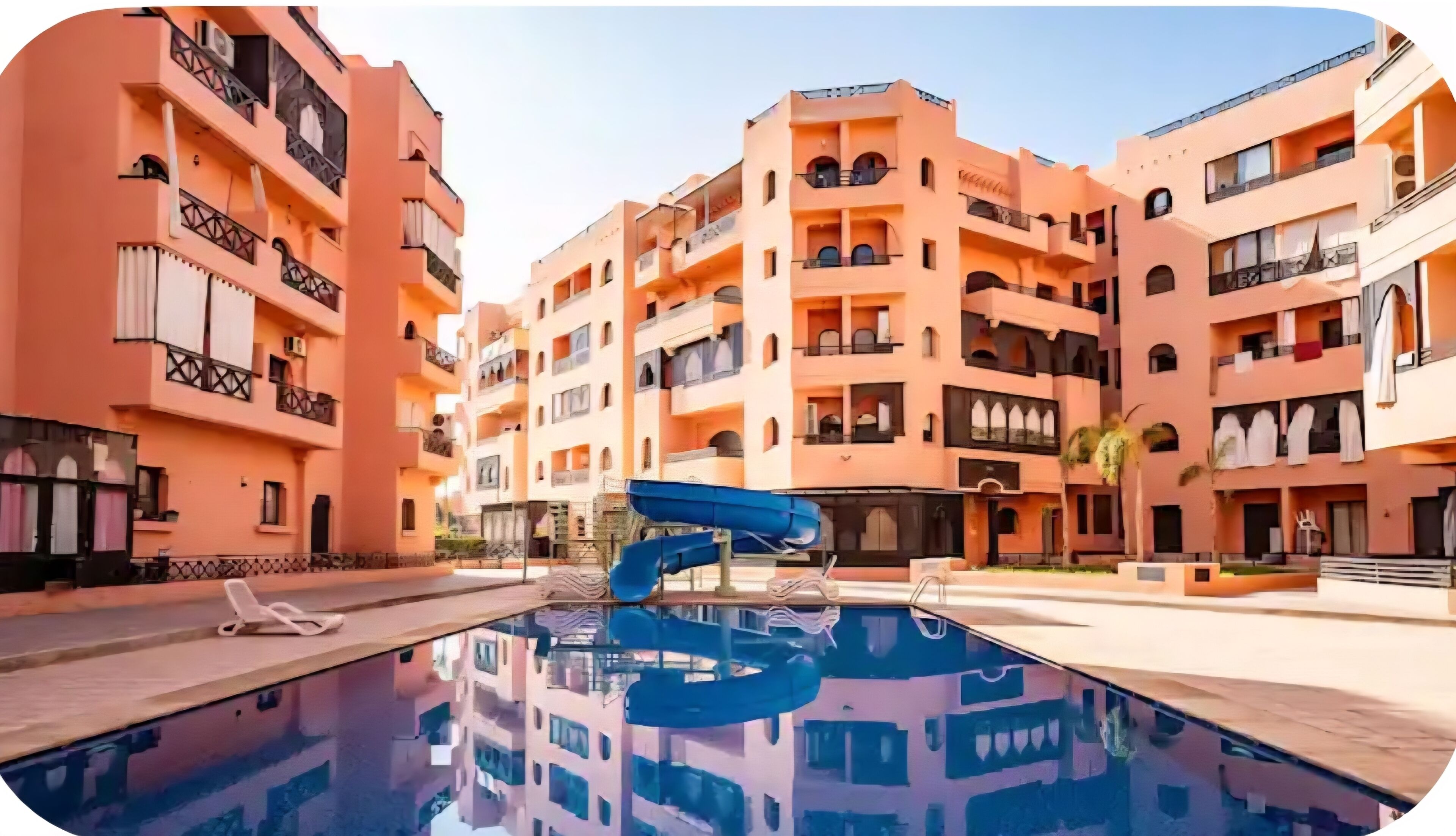 Ein mehrstöckiges Hotel mit Balkonen, einem Swimmingpool mit blauer Rutsche und blauem Himmel.