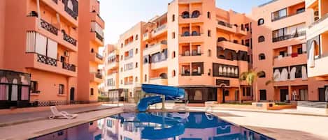 A multi-story hotel with balconies, a swimming pool with a blue slide, and a clear sky.