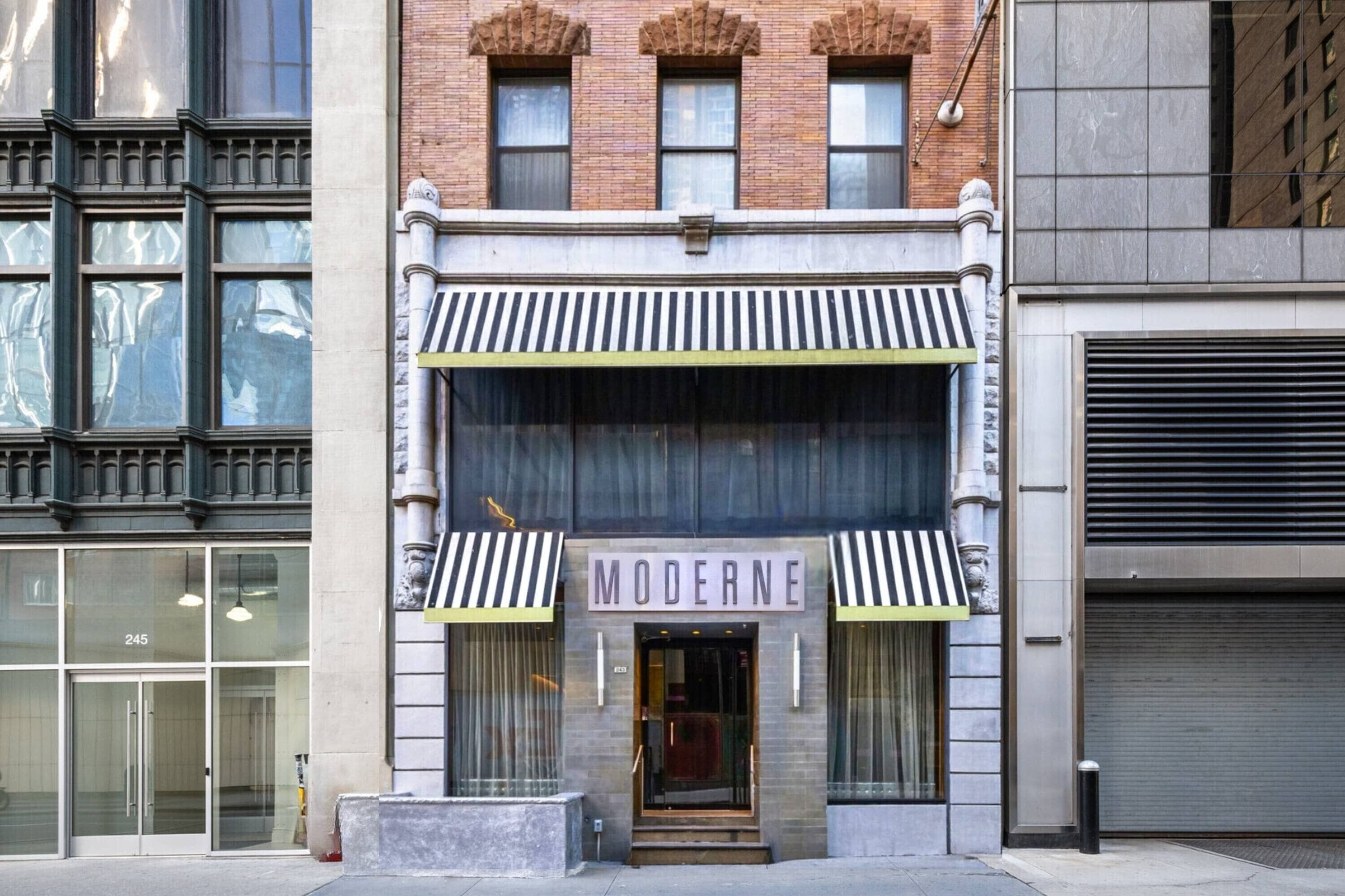 A building with a storefront labeled 'MODERNE' featuring a classic architectural style with a brick facade, large windows, and a striped awning.