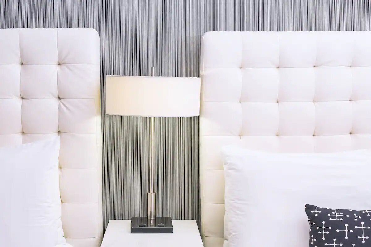 Una habitación con dos cabeceros blancos con capitoné, una lámpara de pie y un cojín estampado.