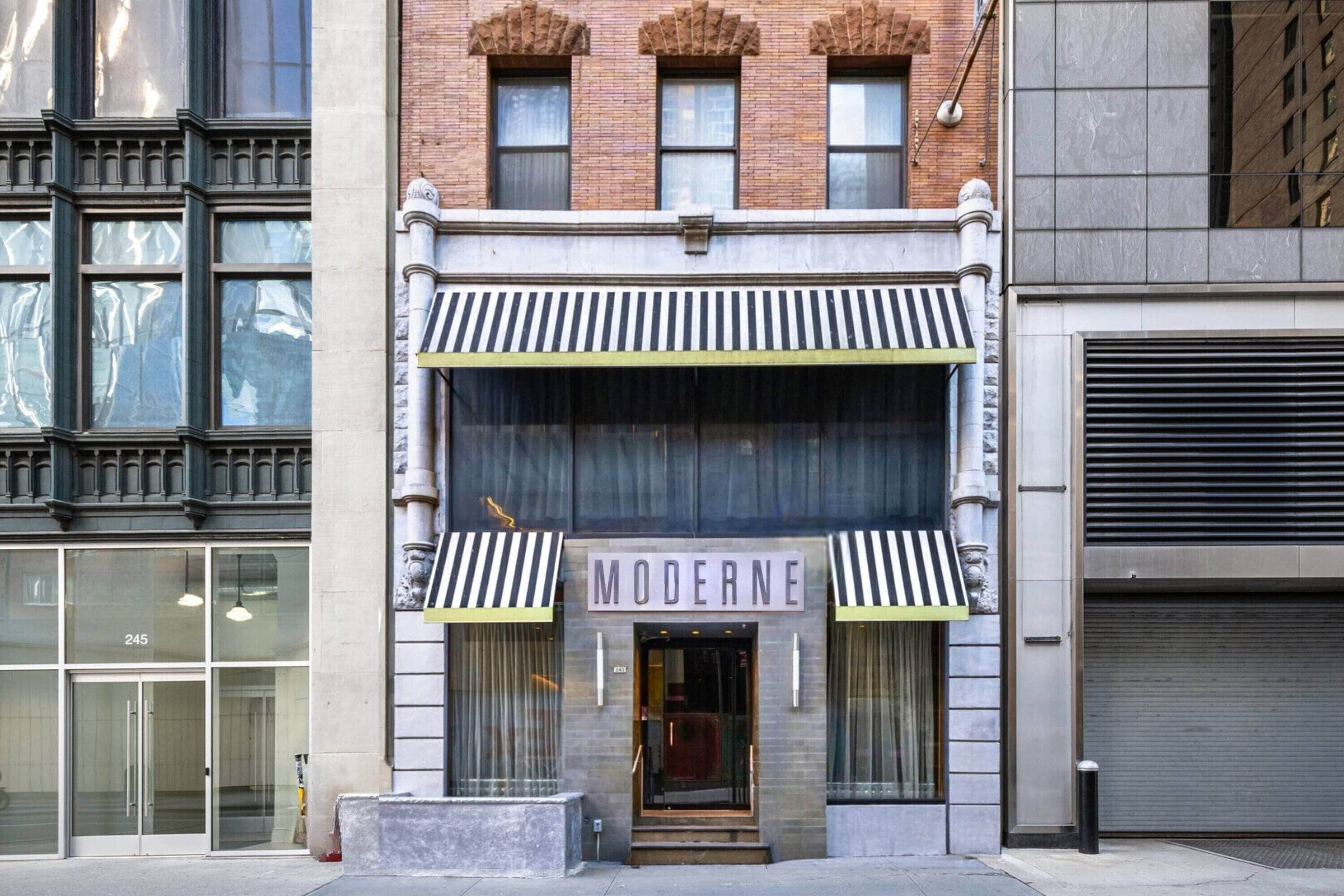 A building with a storefront labeled 'MODERNE' featuring a classic architectural style with a brick facade, large windows, and a striped awning.