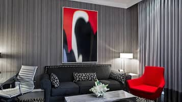 A modern hotel room with a dark grey sofa, a red armchair, a black and white patterned coffee table, and a large abstract painting on the wall.