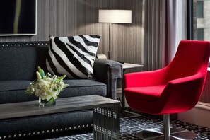 A modern hotel room with a dark sofa, a red armchair, a zebra-patterned pillow, a small table with a vase of flowers, and a lamp. - Moderne | Junior Suite | Midtown Location | 24/7 Guest Lounge | 2 Units (New York)