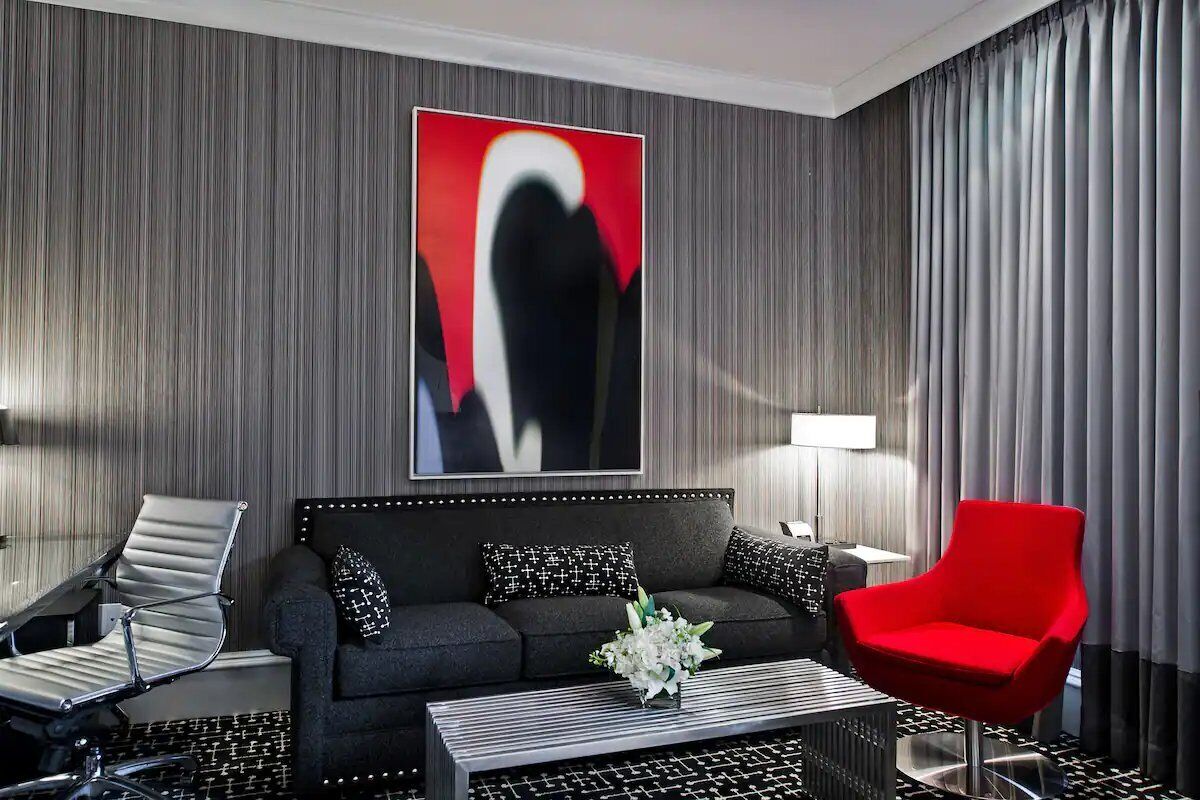 Une chambre d’hôtel moderne avec un canapé gris foncé, un fauteuil rouge, une table basse à motifs noir et blanc et un grand tableau abstrait accroché au mur.