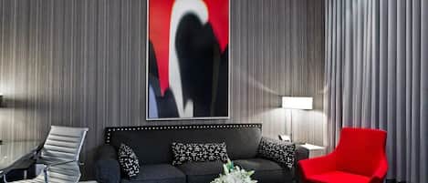 Une chambre d’hôtel moderne avec un canapé gris foncé, un fauteuil rouge, une table basse à motifs noir et blanc et un grand tableau abstrait accroché au mur.