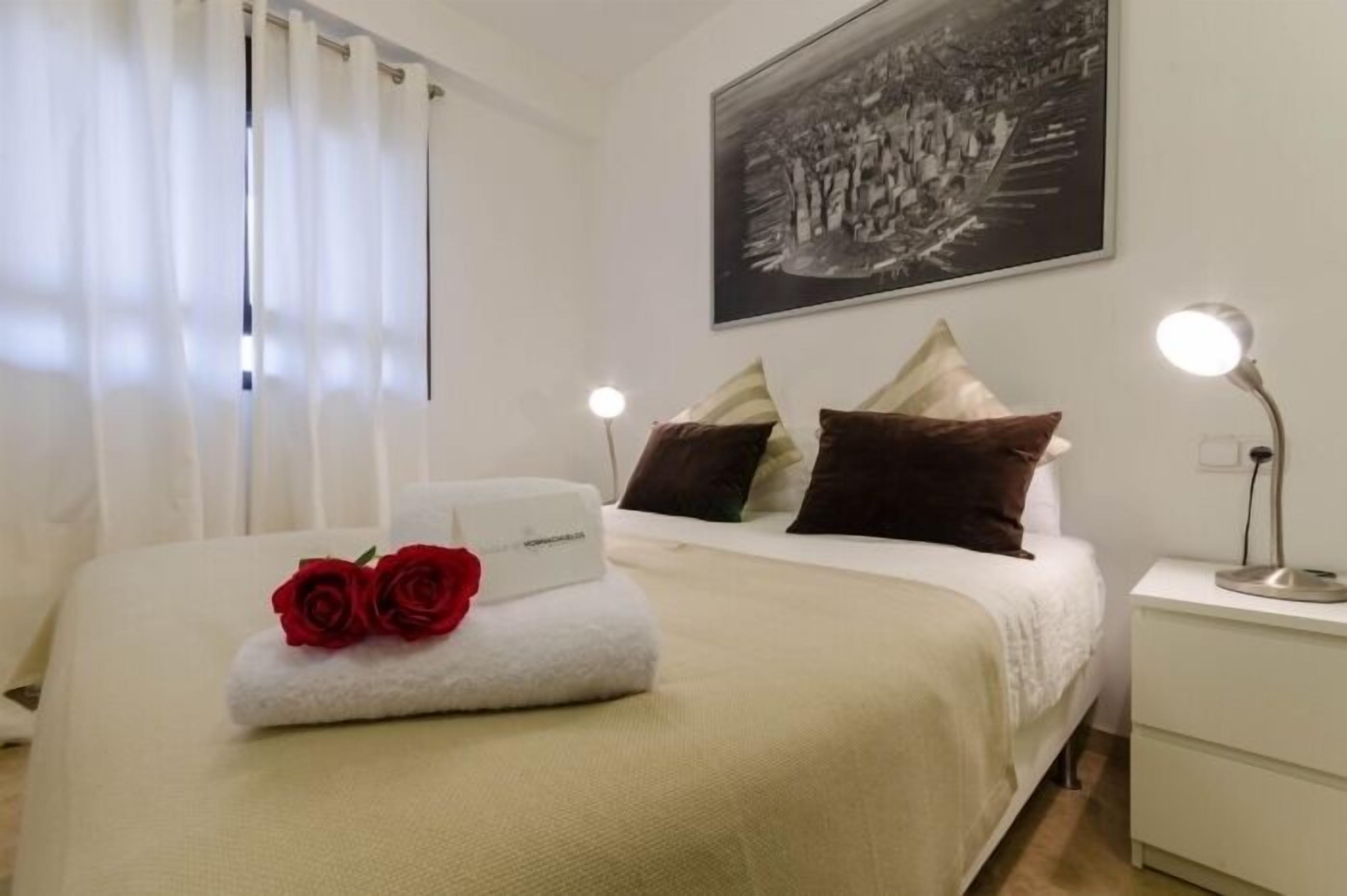 A neatly made bed with a white comforter, two pillows, and a folded towel with red roses. There is a nightstand with a lamp, a framed picture on the wall, and a window with white curtains.