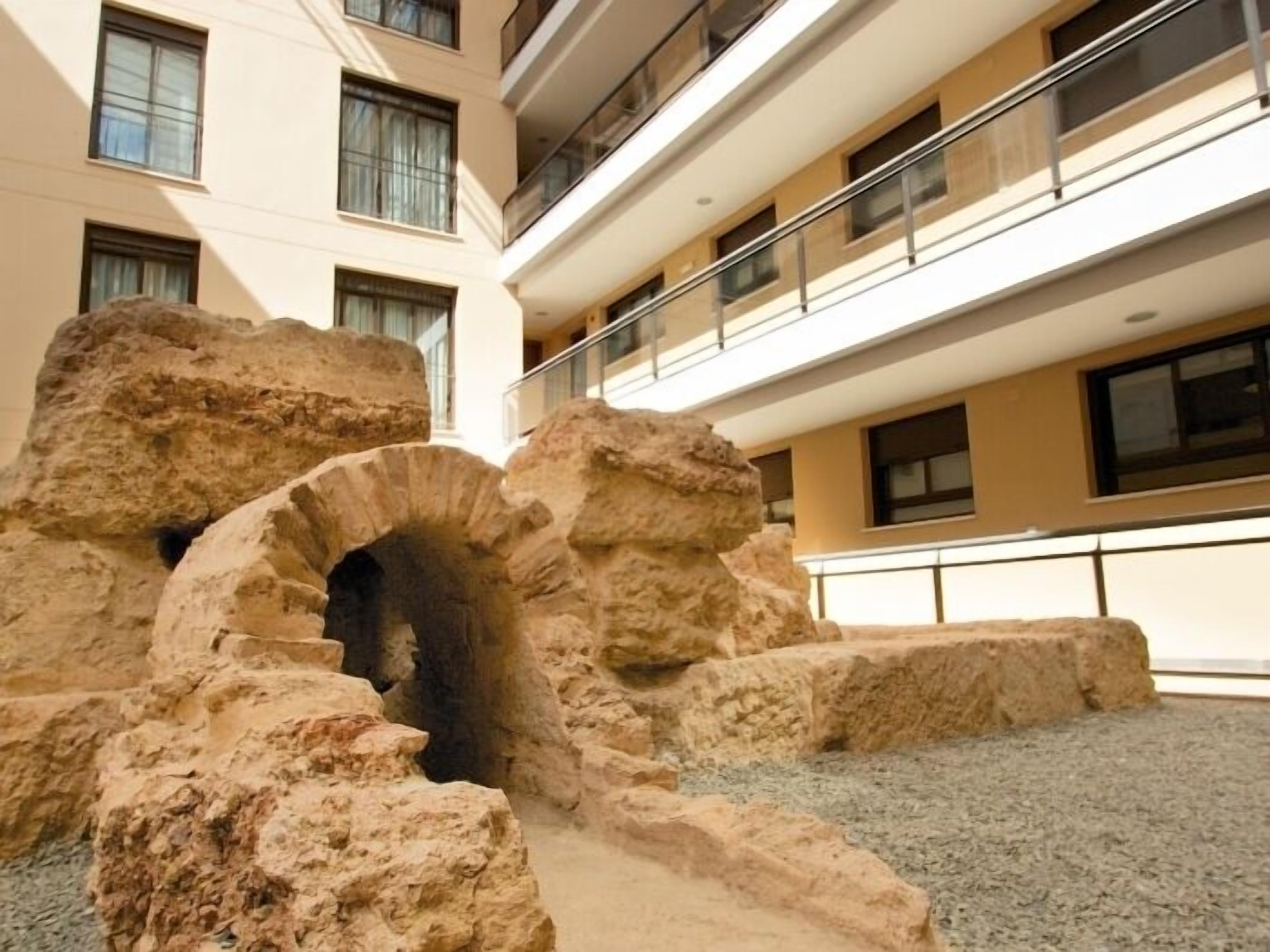 A building with a modern architectural style, featuring a multi-level balcony with railings, large stone structures, and a gravel ground.