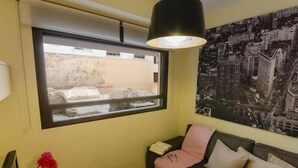 Ein Zimmer mit Fenster, einer Couch, einem kleinen Tisch mit einer Vase und einem Stadtbild an der Wand.